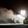 A nighttime construction or industrial scene illuminated by bright work lights, showing a yellow Caterpillar (CAT) compact track loader surrounded by dense white smoke or steam. Several workers are visible as silhouettes standing near the machinery amid the billowing clouds. The dramatic lighting and smoke create a high-contrast, atmospheric composition.