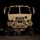 A large tan/desert-colored military tactical truck (consistent with a HEMTT or LMTV-type vehicle) is photographed head-on at night. Several soldiers in military uniforms are lying prone on the ground beneath the front of the vehicle, appearing to conduct an under-vehicle inspection or training exercise. The scene is illuminated by the vehicle's lights and ambient base lighting in an otherwise dark environment.