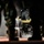 A CAT (Caterpillar) skid steer loader, model W360B, is being operated at night by a person wearing a respirator/gas mask. The scene is framed by silhouetted figures in camouflage military uniforms in the foreground. Dust or smoke is visible near the ground around the machine, suggesting active earthmoving or demolition work.