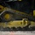 A close-up photograph of a yellow heavy construction machine's rubber track system, likely a compact track loader or similar equipment. The image captures fine mechanical detail of the undercarriage, including drive sprockets, roller wheels, and rubber track pads. Dust or smoke is visible near the ground, suggesting the machine is in active operation on a gravel or debris-covered surface.