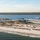 Six U.S. Navy Blue Angels F/A-18 Hornet jets fly in tight delta formation over a coastal barrier island, trailing white smoke streams. The aircraft are flying low over a sandy beach and inlet area, with a forested mainland visible in the background. The image appears to be taken from an aerial vantage point, likely from a chase aircraft or elevated position.