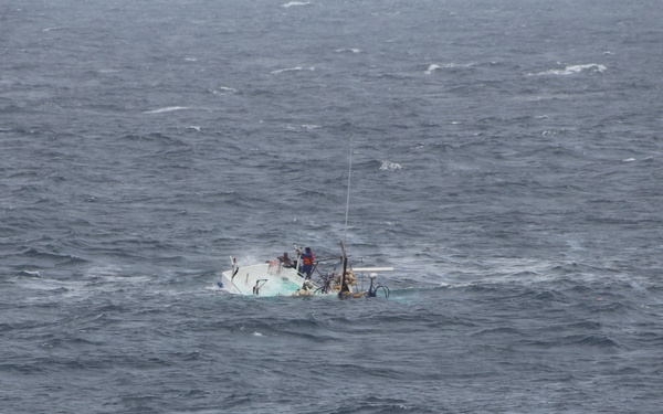 A small vessel appears to be capsizing or severely foundering in rough open ocean conditions. The boat is nearly submerged with only the superstructure, mast, and some rigging visible above the waterline. At least one person in what appears to be an orange survival suit is visible on or near the stricken vessel.
