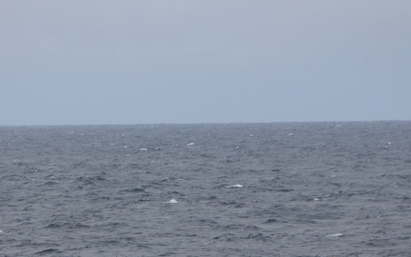 A vessel is capsizing or sinking in rough open ocean conditions, listing heavily to one side with its superstructure partially submerged. A red rigid inflatable boat (RIB), consistent with a Coast Guard or rescue vessel, is positioned nearby, apparently conducting a rescue or response operation. Sea conditions appear rough with choppy waves and overcast skies.