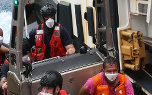 A maritime rescue operation is underway on what appears to be a U.S. Coast Guard vessel. Several personnel in red life vests, helmets, and face masks are attending to a civilian survivor wearing an orange life vest and a pink shirt. The rescued individual appears to have a bandaged hand and is seated on the boat deck.
