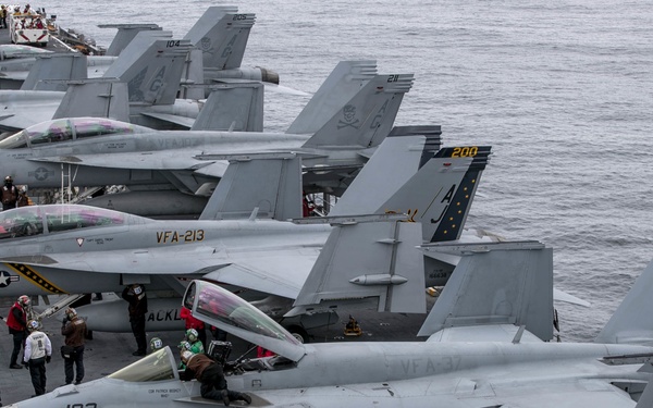 The image shows the flight deck of a U.S. Navy aircraft carrier crowded with multiple F/A-18 Super Hornet fighter jets from several squadrons. Deck crew in color-coded jerseys are performing pre-flight, maintenance, and aircraft handling operations. The carrier appears to be at sea, with open ocean visible in the background.