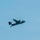 A United States Navy turboprop aircraft is photographed in flight against a clear blue sky. The aircraft appears to be a C-2 Greyhound, identifiable by its twin engines, high-wing configuration, and distinctive tail markings. The aircraft displays orange/yellow tail markings and a US Navy star insignia on the fuselage.