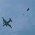 A military turboprop aircraft is captured in flight against a partly cloudy blue sky, with a paratrooper visible to the right having just exited the aircraft. The paratrooper's parachute appears to be in the early stages of deployment, with a small drogue or pilot chute visible above the jumper. The aircraft displays visible US military markings (star insignia) on its underside.