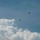 The image shows a military or cargo aircraft in flight with at least one parachutist visible in the sky, having already deployed their parachute. The scene is set against a partly cloudy blue sky with large cumulus clouds in the lower portion of the frame. Both the aircraft and parachutist appear as dark silhouettes at high altitude.