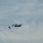 A twin-engine turboprop aircraft, consistent with a C-2 Greyhound or similar carrier onboard delivery (COD) aircraft, is captured mid-flight performing an airdrop operation. Two objects — appearing to be cargo packages — have been released from the aircraft and are descending with parachutes deployed. The image is taken against an overcast sky with diffuse cloud cover.