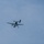 A military turboprop transport aircraft is captured mid-flight with a paratrooper in the process of exiting the aircraft. The parachute appears to be in the early deployment stage, still partially collapsed above the jumper. The image was taken from a ground-level perspective against an overcast sky.