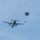 A military transport aircraft is captured mid-flight with a paratrooper who has just exited the aircraft and deployed a parachute. The parachute appears to be a round military-style canopy in an olive/dark green color. The scene takes place against a pale blue, slightly overcast sky.