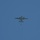 A twin-engine turboprop military aircraft is photographed from below against a clear blue sky. A small object or figure is visible falling or suspended below the aircraft, with a second object visible further below, suggesting an aerial drop operation in progress.