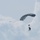 A parachutist is descending through a partly cloudy sky using a dark gray ram-air parachute canopy. The jumper appears to be in full descent with control toggles visible, and a small aircraft can be seen in the background to the left. The image captures what appears to be a military or sport skydiving operation.