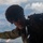 A military service member in full tactical gear is leaning out of what appears to be an aircraft, likely preparing for or conducting an airborne operation. The individual is wearing a combat helmet, protective eyewear, multicam/OCP patterned uniform, and a parachute harness or tactical vest. The sky with scattered clouds is visible in the background, indicating significant altitude.