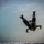 A military or tactical skydiver is captured in freefall immediately after exiting an aircraft, visible at the bottom edge of the frame. The jumper is in a tumbling, head-down orientation over a coastal area with teal-blue water and a sandy shoreline visible far below. The image is taken from the aircraft's perspective looking down.