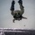 A military parachutist in full combat gear is captured mid-freefall in an inverted (head-down) position above a large body of water. The individual is wearing camouflage uniform with a parachute rig on their back and fins attached to their boots, indicating a combat water insertion (HALO/HAHO) operation. The image is taken from above, likely from the aircraft the jumper just exited, with a structure visible at the bottom edge of the frame.