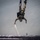A military skydiver in full camouflage uniform and tactical gear is captured in a head-down freefall position over a large body of water. The jumper appears to be in a controlled vertical dive, with a pilot chute or drogue streamer visible trailing from their body. A structure, possibly a bridge or pier, is partially visible at the bottom edge of the frame.