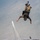 A military skydiver in full camouflage uniform and tactical gear is captured in a head-down freefall position over a large body of water. The jumper is inverted with feet pointing upward, wearing a parachute rig and harness system. A small boat is visible far below on the water's surface, leaving a white wake trail.