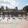 A group of military personnel in camouflage uniforms are standing in formation on a paved plaza, each holding red and yellow guidon flags with visible numbers. They appear to be at a U.S. Marine Corps base, likely Marine Corps Recruit Depot (MCRD) San Diego, based on the architectural style and setting. The formation is positioned in front of a memorial or monument with a bronze plaque.