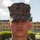 A young male service member in U.S. Marine Corps MARPAT woodland digital camouflage uniform and cover (cap) with the Eagle, Globe, and Anchor emblem visible on the front. He is posed facing the camera in a formal portrait-style photograph taken outdoors. The background shows a military installation with trimmed hedges and a tan/beige building.