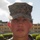 A close-up portrait of a young male service member wearing United States Marine Corps MARPAT (Marine Pattern) digital camouflage utilities and cover (cap). He is wearing a dog tag chain visible at the collar and has a serious, composed expression. The background shows trimmed hedges and a building consistent with a military base environment.
