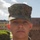 A close-up portrait of a young male service member wearing U.S. Marine Corps MARPAT (Marine Pattern) digital camouflage utilities and cover (cap). The subject is photographed outdoors against a partly cloudy sky with institutional buildings and trimmed hedges visible in the background. The image appears to be an official or semi-official military portrait.