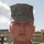 A close-up portrait of a male service member wearing U.S. Marine Corps MARPAT woodland digital camouflage uniform and cover (cap). The subject is standing at attention or in a formal pose outdoors, with a military installation building visible in the background. The image appears to be an official or semi-official military portrait photograph.