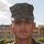 A young male service member in U.S. Marine Corps MARPAT (woodland digital camouflage) uniform and cover (cap) is photographed in a portrait-style shot. He is wearing a dog tag chain visible at the neckline. The background shows a military installation or base facility with manicured hedges and a multi-story building.