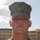 A young male service member in U.S. Marine Corps MARPAT woodland digital camouflage uniform and cover (cap) is photographed in a formal portrait-style image. He is standing at attention with a neutral expression against an outdoor background featuring a building and partly cloudy sky. The Eagle, Globe, and Anchor emblem is visible on his cover.