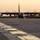 Sweden and U.S. Strengthen Air Capabilities During Swedish-led Bilateral Exercise A large four-engine turboprop military transport aircraft, consistent with a C-130 Hercules variant, is taxiing on a runway or taxiway at a military airfield during golden hour (sunrise or sunset). All four propellers are spinning, visible as motion-blurred discs. The low-angle shot from runway level creates a dramatic perspective with airfield buildings and trees visible in the background.