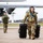 Sweden and U.S. Strengthen Air Capabilities During Swedish-led Bilateral Exercise Military personnel in OCP (Operational Camouflage Pattern) uniforms are walking across an airfield tarmac near a large military aircraft. The foreground figure is carrying two black hard-shell equipment cases while wearing hearing protection headsets. The scene appears to be a deployment or equipment offload operation at a military air base.
