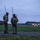Sweden and U.S. Strengthen Air Capabilities During Swedish-led Bilateral Exercise Two military personnel in multicam camouflage uniforms stand on the edge of an airstrip at dusk or dawn, each equipped with tall radio antennas and tactical backpacks. They are observing a military transport aircraft — likely a C-130 Hercules — taxiing or landing on the runway in the background. The setting appears to be a forward or austere airfield surrounded by dense tree lines.
