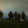 Sweden and U.S. Strengthen Air Capabilities During Swedish-led Bilateral Exercise A group of approximately five to six military or tactical personnel are standing in an open field at night. They are equipped with full tactical gear including helmets with night vision devices (NVDs) that emit a faint blue-green glow. The scene appears to be a nighttime military operation or training exercise, with distant lights visible on the horizon.