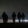 Sweden and U.S. Strengthen Air Capabilities During Swedish-led Bilateral Exercise A nighttime photograph showing five silhouetted figures wearing helmets and tactical gear standing in a line facing away from the camera. Behind them, light trails from passing vehicles create streaks of red, white, and orange across the frame, suggesting a long-exposure photograph taken near a roadway. The dark sky and low ambient light render the figures almost entirely in silhouette.