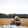Sweden and U.S. Strengthen Air Capabilities During Swedish-led Bilateral Exercise A military paratrooper in full combat gear is kneeling on an open field, gathering and packing a deployed parachute after a landing. The soldier is wearing MultiCam (OCP) camouflage uniform, a tactical helmet with night vision mount, hearing protection, and carries a rucksack. The setting appears to be a drop zone in a flat, grassy field with a treeline visible in the background.