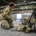 Sweden and U.S. Strengthen Air Capabilities During Swedish-led Bilateral Exercise A soldier in multicam camouflage uniform is kneeling on the floor of a large military hangar, appearing to organize or inspect equipment. Several rifles are leaned against a military vehicle tire alongside tactical gear including a helmet and backpack. At least one other soldier is visible in the background near an open hangar door.