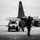 Sweden and U.S. Strengthen Air Capabilities During Swedish-led Bilateral Exercise A black-and-white photograph taken from inside a hangar looking outward onto a military airfield tarmac. Armed personnel in tactical gear are walking toward the hangar entrance, accompanied by a light tactical vehicle, with a large military transport aircraft (consistent with a C-130 Hercules) visible in the background. The composition creates a dramatic silhouette effect with strong contrast between the dark hangar frame and the overcast exterior.