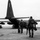 Sweden and U.S. Strengthen Air Capabilities During Swedish-led Bilateral Exercise A black-and-white photograph showing military personnel in camouflage uniforms and tactical gear approaching a USAF C-130 Hercules transport aircraft on a tarmac. Two soldiers are prominently visible in the foreground walking toward the aircraft's rear ramp, while a group of additional personnel is visible to the left near the aircraft. The tail markings indicate this is a United States Air Force aircraft.