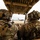 Sweden and U.S. Strengthen Air Capabilities During Swedish-led Bilateral Exercise Two military personnel in full combat gear are seated inside a tactical vehicle, viewed from behind, on what appears to be an airfield tarmac. They are wearing multicam camouflage uniforms, tactical helmets with communications equipment, and body armor. A military transport aircraft, consistent with a C-130 Hercules, is visible in the background on the runway.