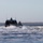 Sweden and U.S. Strengthen Maritime Capabilities During Swedish-led Bilateral Exercise Two military-style patrol or special operations boats are navigating through choppy coastal waters, with the lead vessel displaying an American flag. The boats appear to be moving at speed, generating significant wake and white water. Wind turbines and a treeline are visible on the distant shoreline in the background.