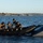 Sweden and U.S. Strengthen Maritime Capabilities During Swedish-led Bilateral Exercise The image shows a group of military or special operations personnel on a rigid inflatable boat (RIB) alongside a larger gray vessel on open water. Several individuals with obscured faces are handling what appears to be a cylindrical piece of equipment or weapon system in the inflatable boat. A fifth person stands on the larger vessel observing the activity, with a rocky coastline visible in the background.