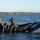 Sweden and U.S. Strengthen Air Capabilities During Swedish-led Bilateral Exercise Military or special operations personnel are conducting a maritime exercise involving a rigid inflatable boat (RIB) alongside a larger angular stealth-style vessel. Several personnel in tactical gear are working on or around the RIB, appearing to handle equipment near the waterline. The setting appears to be a Scandinavian archipelago based on the rocky, forested shoreline visible in the background.