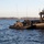 Sweden and U.S. Strengthen Maritime Capabilities During Swedish-led Bilateral Exercise The image shows Swedish military personnel operating on open water using two vessels: a rigid inflatable boat (RIB) and a larger military patrol/assault craft flying the Swedish flag. Personnel in military attire and face coverings are visible aboard both vessels, suggesting a training exercise or operational activity. The scene takes place on a calm inland waterway or coastal area with a tree-lined shore visible in the background.