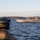 Sweden and U.S. Strengthen Maritime Capabilities During Swedish-led Bilateral Exercise A military or law enforcement personnel in tactical gear and helmet observes a stealth-style vessel on open water from a nearby boat. The vessel in the background has an angular, low-profile design consistent with special operations or prototype watercraft, and flies an American flag. The scene appears to take place in a coastal or large inland waterway with industrial or institutional structures visible on the distant shoreline.