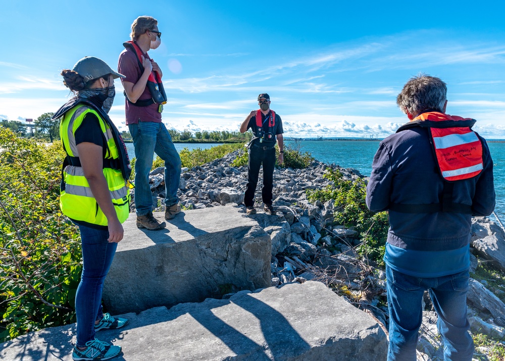 Four individuals are gathered on a rocky waterfront structure, appearing to conduct a site inspection or safety briefing. Three of the four people are wearing personal flotation devices (PFDs) or life vests, and all are wearing face masks, suggesting this was taken during the COVID-19 pandemic period. The setting appears to be a deteriorating concrete and rock breakwater or jetty along a large body of water.