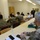 Guard medical team supports Czech partners A meeting or briefing is taking place in a conference room between military personnel in camouflage uniforms and civilians in white shirts and business attire. Several participants are wearing face masks, suggesting this occurred during the COVID-19 pandemic period. The setting appears to be a formal but modest meeting room with yellow-clothed tables arranged in a U-shape or classroom style.