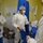 Guard medical team supports Czech partners A group of healthcare workers are donning personal protective equipment (PPE) in what appears to be a hospital or medical facility preparation area. The individuals are wearing white protective suits, hair nets, face masks, and are handling blue protective garments. The setting suggests a high-infection-risk environment requiring full PPE protocols.
