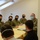 A group of U.S. Army personnel in camouflage uniforms and face masks are seated around a table with a yellow checkered tablecloth, engaged in what appears to be a meeting or briefing. They are facing two individuals whose backs are to the camera. The setting appears to be an indoor room with slanted ceiling skylights, suggesting a European-style building.