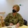 A U.S. Air Force service member wearing OCP (Operational Camouflage Pattern) uniform and a black face mask is seated at a table, taking notes in a notebook. The name tape reads a partially visible surname and the chest tape reads 'U.S. AIR FORCE.' The individual appears to be attending a meeting or briefing in an indoor setting.