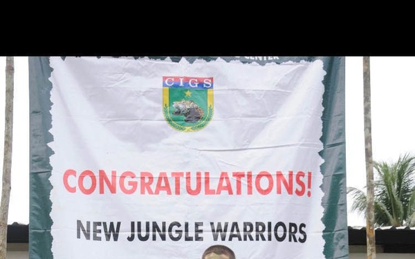 A soldier in full camouflage uniform and face paint stands in front of a congratulatory banner marking completion of jungle warfare training at CIGS (Centro de Instrução de Guerra na Selva - Jungle Warfare Training Center). The soldier is holding an assault rifle and wearing full combat gear. The banner reads 'CONGRATULATIONS! NEW JUNGLE WARRIORS - NOW YOU ARE ONE OF US!'
